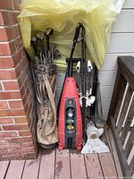 Wide view of golf clubs, red power washer, ski equipment, and ski pole under yellow plastic covering on deck next to brick wall.