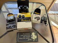 Top-down view of the lot with three binoculars (one full-size Bushnell, two small ones, one boxed) and a wooden sign with a John Muir quote.