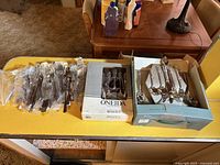 Silverware sets laid out on a table including a boxed Oneida set and loose pieces of silverware, plus another boxed Cambridge silverware set with spoons, forks, knives.