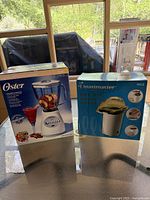 Photo showing boxed Oster blender and Toastmaster hot air popper side by side on table, natural light from window