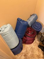 Six sleeping bags rolled and positioned upright on carpeted floor in a corner, showing variety of colors and storage conditions.
