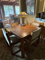 Square wood kitchen table with four matching chairs and decorative lamp on top, showing the setting in a wood-paneled room.