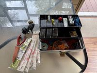 Top-down view of open tacklebox with compartments holding fishing tackle including lines, lures, bobbers, and other fishing accessories, along with fishing hooks and a red handled tool beside it.