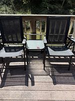 Two black mesh fabric fold-up lounge chairs positioned outdoors on a wooden deck with a small glass-top side table between them.