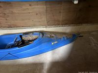 Side view of blue Water Quest Aruba kayak in a wooden and concrete indoor space, showing heavy dirt and mud accumulation on and around cockpit area.