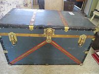 Front side view of the antique trunk showing metal latch, side clasps, and red diagonal striping on blue body.