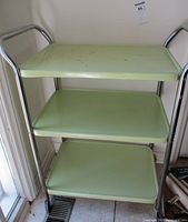 Front view of the vintage cart showing three green metal shelves with chrome frame and handles.