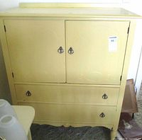 Front view of vintage highboy chest with two doors above two large drawers, painted pale yellow, showing metal ring drawer pulls and subtle decorative carving.