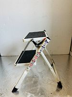 Full view of the white metal two step ladder with black rubber non-slip steps, side profile showing ladder open and standing.