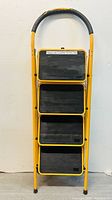 Front view of 4-step foldable ladder showing black textured anti-slip steps and yellow metal frame with black cushioned top handle.