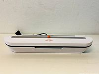 Closed view of white and gray rectangular food vacuum sealer with orange button on top and power cord attached.