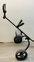 Side view of the black golf push/pull cart showing large rear wheel, smaller front wheel, handle, and basket.