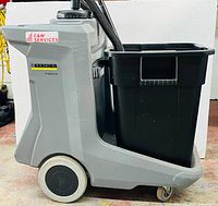 Side view showing gray plastic cart body, integrated black trash bin, large rear wheel and front caster, vacuum hose stowed on top