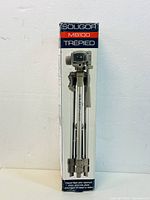 Front of the original Soligor MB100 tripod box showing the tripod stand with head and folded legs in silver color.