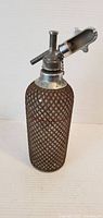 Front and top angle of the Old Faithful wire mesh glass syphon seltzer bottle showing metal spout lever and mesh details