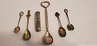 Photo showing six souvenir collectible spoons and one small decorative keychain or charm with colored inlay. Various sizes and designs including an integrated bottle opener handle on one spoon.