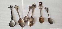 Photo showing six souvenir spoons of varying designs and conditions arranged side by side on a white background.