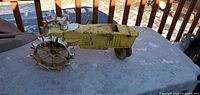 Side view showing the overall shape of the heavy metal yellow tractor sprinkler with rust and serrated wheels on a table outdoors.