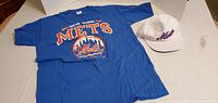 Blue New York Mets T-shirt and white Mets hat laid flat on a white surface.