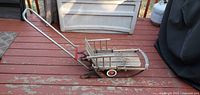 Full view of vintage wooden sled showing wooden seat and backrest, metal runners, wheels with red rims, and pushing handle.
