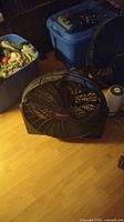 Black Honeywell Big Fan positioned on wooden floor with blue plastic bin and miscellaneous items in background.