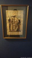 Framed Egyptian painting on papyrus showing a traditional scene with multiple figures, mounted on a green mat and enclosed in a wooden frame with glass.