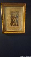 Framed Egyptian painting on papyrus hung on a dark wall showing two figures in traditional attire with decorative gold frame