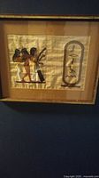 Framed painting on papyrus showing three Egyptian style figures next to a cartouche with hieroglyphics.