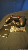 Photo of a black Gamesir Gamenote T3 wireless game controller with red-tipped analog sticks, a USB charging cable coiled in front, a user manual, and a small sealed plastic bag containing additional accessories, all placed on a dark-colored fabric surface.