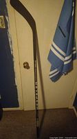 Full view of the black Hespeler Doug Gilmore hockey stick leaning against a door, showing the blade and shaft with white lettering.