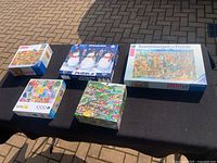 Five boxed 1000-piece puzzles arranged on a black table with a brick ground background. Includes Ravensburger, snowmen, countryside scene, and other puzzles.