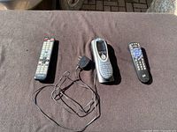 Three remote controls placed on a fabric surface outdoors in sunlight. Logitech Harmony remote with charging adapter in the middle, other two remotes flanking it.