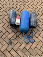 Three blue and dark colored cylindrical boat fenders alongside a red and white crab float on a brick pavement.