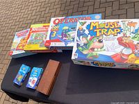 Photo displaying 5 boxed board games including Mouse Trap, Operation Toy, Guess Who?, 3D Snake Serpents, and an additional Mousetrap game box along with two playing card decks and a score pad on black tablecloth.