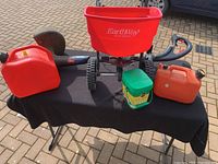 Full lot showing the weed trimmer, two gas cans, the red EarthWay fertilizer spreader, and Miracle-Gro fertilizer container on a black table.