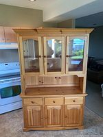 Front view of pine hutch showing upper glass cabinets, open shelf, and lower storage with three doors and two drawers. Kitchen stove visible on the left side.