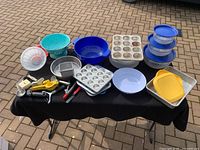 Overview of all kitchen items on outdoor table, showing plastic colanders, bowls, baking pans, Tupperware stack, muffin tins, and assorted utensils.