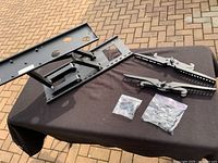 Overview photo of the black metal mounting plate, two adjustable arms and two bags of assorted bolts and mounting hardware arranged on a black cloth over stone tiles outside.