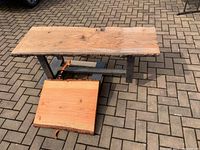 Photo of the full live edge wood table with steel base positioned on outdoor paved surface, smaller untreated wood piece beneath table top for size and wood comparison.