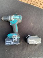 Makita cordless drill with 18V 5.0Ah battery attached and 3.0Ah battery separate on a black surface outdoors.