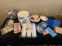 Overview of party supplies showing stacked plates, plastic cups, various plates with colorful designs, cutlery, and small items in plastic bags.