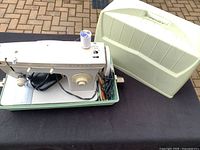 Singer 247 sewing machine seated in its matching green base with power cord and foot pedal wires visible beside it, next to the plastic carrying case.