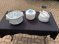 Photo of three kitchen appliances, including a Rival Crock-Pot slow cooker, a white ceramic crock pot with lid, and an electric kettle on a black cloth outdoors on brick flooring.