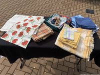 Image showing a black table with an assorted set of fabric tablecloths, placemats, napkins, and wooden napkin rings arranged in stacks.