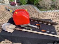 Photo shows shovel, jerry can, car jack, and broom head laid out on a black surface outdoors on stone tiles with garden in background.