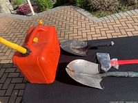 Photo showing red plastic gas can with yellow spout and two shovels including one with broken tip and one with soil on blade plus new axe with red handle wrapped in plastic.