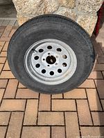 Single trailer tire mounted on steel rim displayed upright against stone wall on brick pavement.