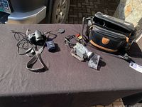 Lot shown on outdoor surface with Lumix still camera, video camera, cables, charger, camera bag shown.