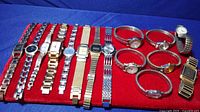 15 assorted women's watches displayed on red cloth, featuring different metal stretch and link bands, round and rectangular faces