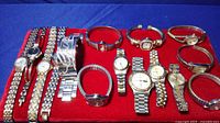 Assortment of 15 women's metal band wristwatches laid out on a red fabric background, showing a variety of face shapes and band styles in silver and gold tones.